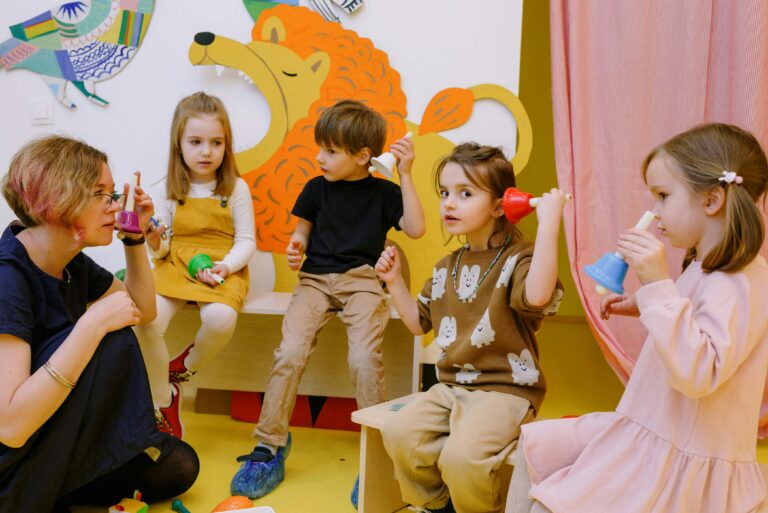 Young children playing musical bells in a colorful classroom setting with a teacher.